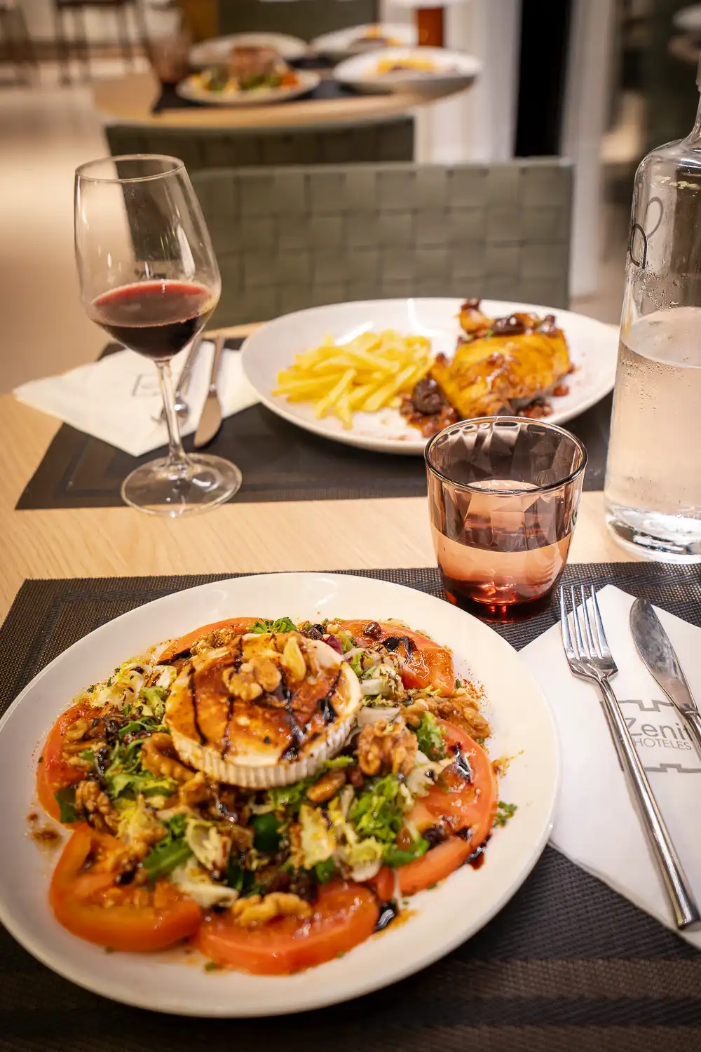 Ensalada de queso de cabra sobre tomate con una copa de vino tinto y pollo con patatas al fondo