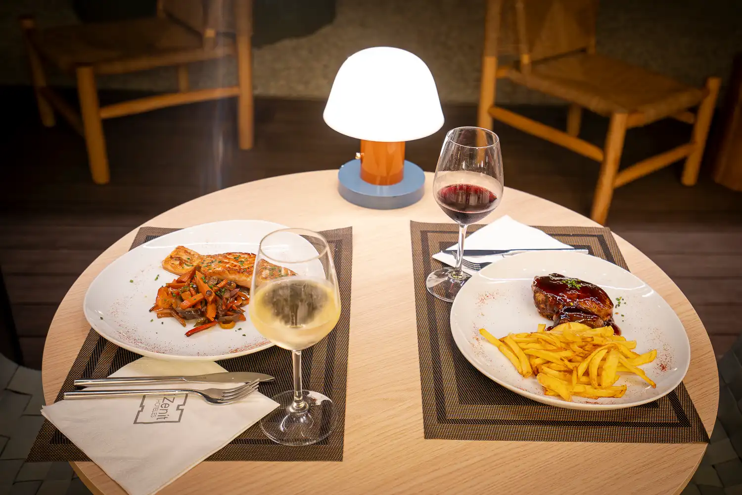 Mesa redonda para dos de noche con una lámpara encendida, salmón con verduras, carne con patatas fritas y copas de vino blanco y tinto