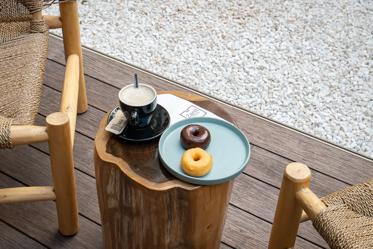 Café y dos donuts sobre un taburete de madera en la tarima del patio, con grava blanca