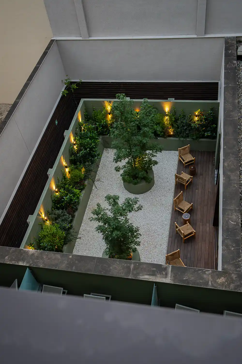 Patio interior visto desde arriba al anochecer, con árboles, suelo de grava, tarima de madera y luces encendidas entre las plantas