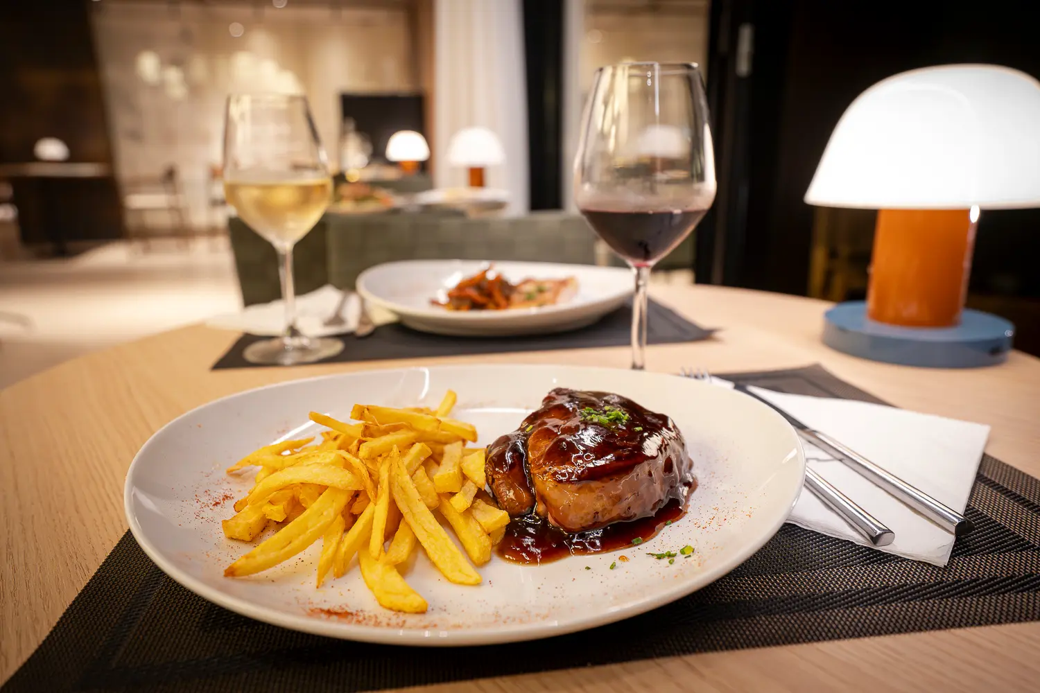 Carne en salsa con patatas fritas, una copa de vino tinto y otra de blanco en una mesa del comedor con lámpara de sobremesa